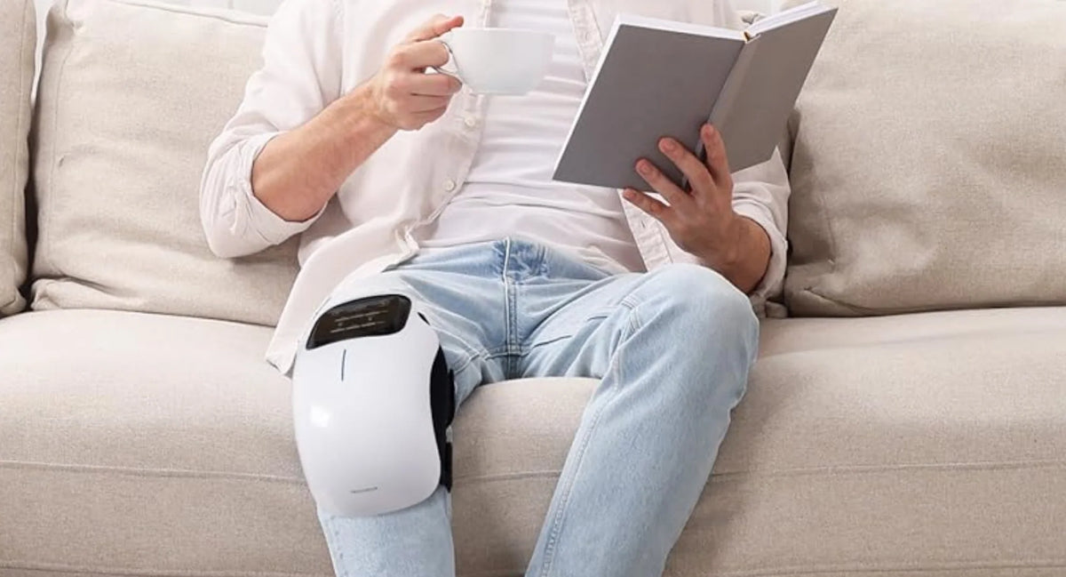 Person sitting on a couch with a knee pain relief massager, holding a book and a mug.